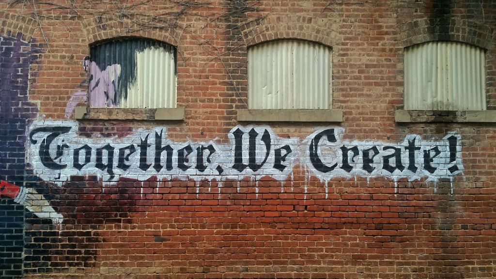 Photo of an old brick wall with graffiti saying, 'Together, We Create!'.